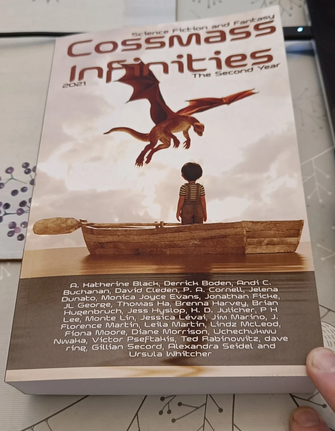 Photo of Cossmass Infinities The Second Year. The cover shows an illustration of a boy standing with his back to us in a rowing boat. Hovering in the air in front of him is a small red dragon.